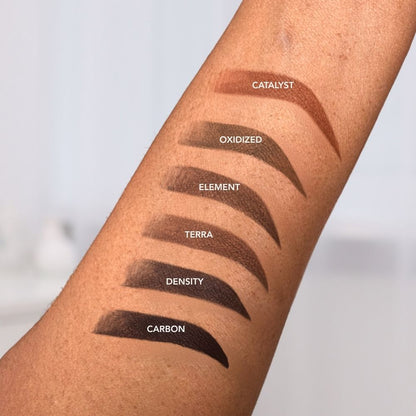 Six INK brow pigment shades swatched on a forearm, in the shades CATALYST, OXIDIZED, ELEMENT, TERRA, DENSITY, and CARBON