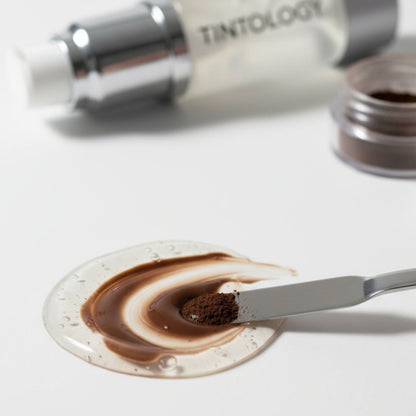 Close-up studio shot of a cosmetic mixing process: a pump of clear Tintology G2 Fuse Activator Gel is blended with a mound of dark brown True Tone Pigment powder using a small metal spatula.