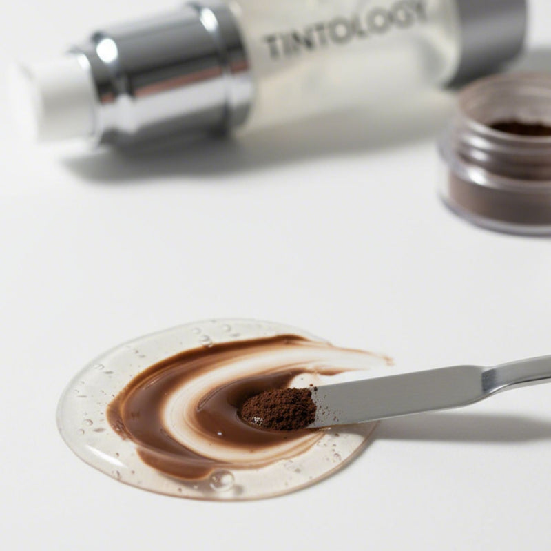 Close-up studio shot of a cosmetic mixing process: a pump of clear Tintology G2 Fuse Activator Gel is blended with a mound of dark brown True Tone Pigment powder using a small metal spatula.